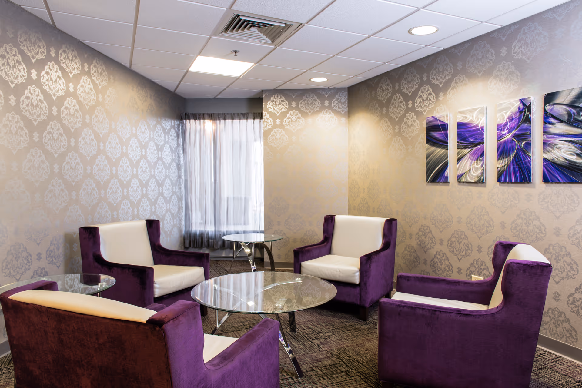 A small sitting area with four purple and white armchairs arranged around a round glass coffee table. The room has patterned wallpaper in a light gray tone, a window with sheer curtains, and four abstract purple and white paintings on the wall.