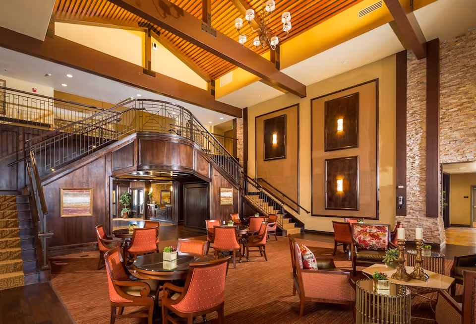 Spacious elegant communal lounge with round tables, red upholstered chairs, a central wooden staircase and high beamed ceiling.