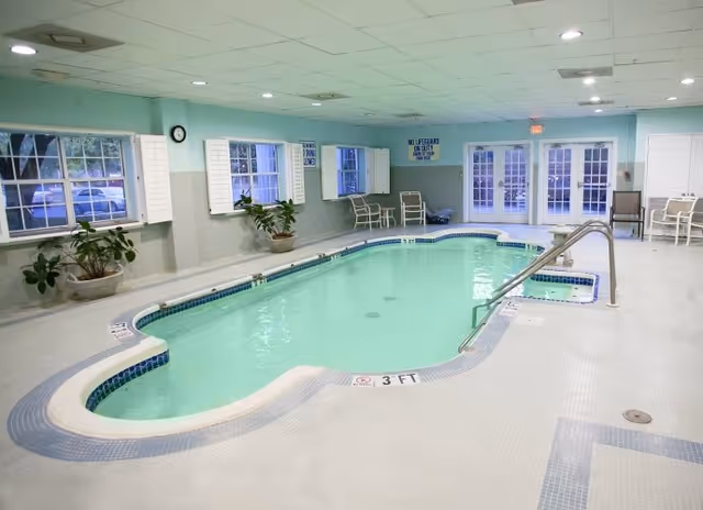 Indoor swimming pool with light blue water surrounded by white tiled floor and light blue walls. Several potted plants and white chairs are placed along the walls. Large windows with white shutters and glass doors are visible in the background. A sign on the wall reads 'NO LIFEGUARD ON DUTY SWIM AT YOUR OWN RISK.'