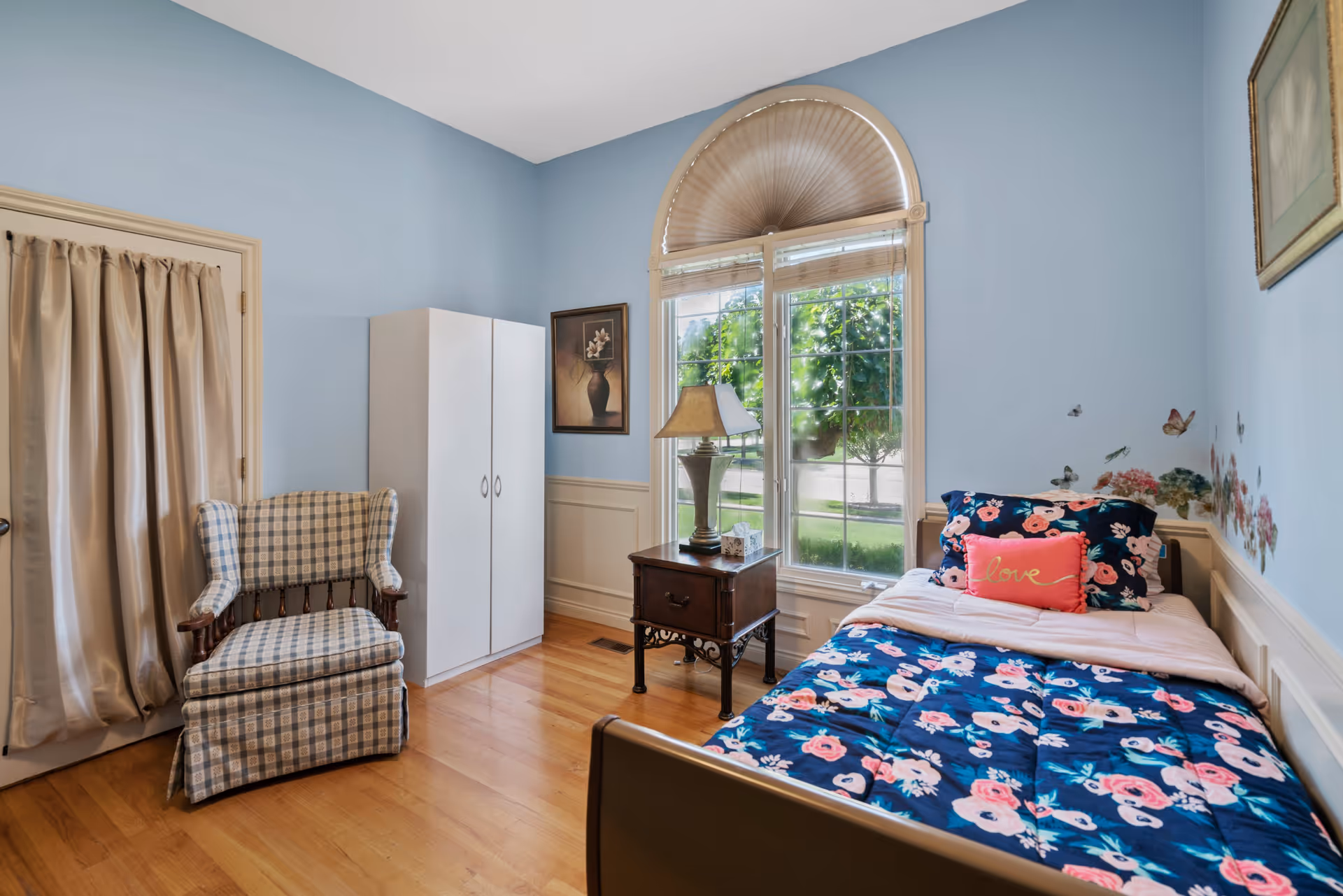 A cozy bedroom with light blue walls and hardwood floors. The room features a single bed with floral bedding and a pink pillow that says 'love'. Next to the bed is a wooden nightstand with a lamp and a tissue box. There is a large window with an arched top and blinds, letting in natural light and showing greenery outside. A white wardrobe and a cushioned armchair with a checkered pattern are also in the room. Two framed pictures hang on the walls.