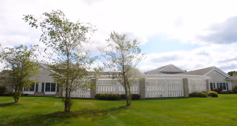 Low single-story brick and siding senior living building with a white fenced patio, green lawn and small trees under a partly cloudy sky.