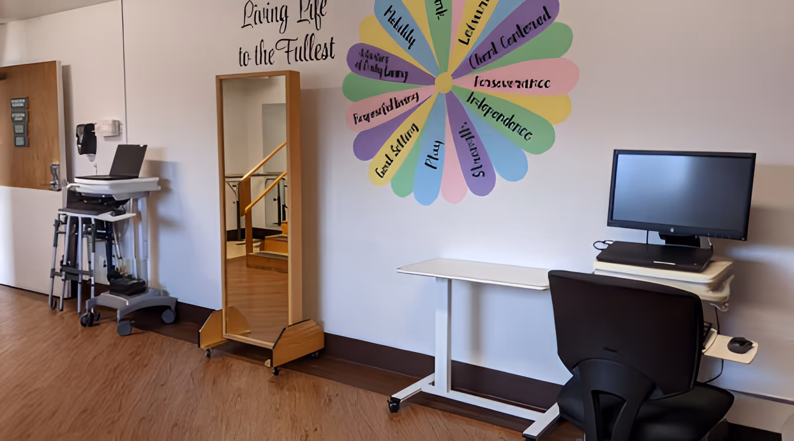 An interior room with a wooden floor, a full-length mirror on wheels, a computer workstation with a monitor and chair, and a colorful wall decoration shaped like a flower with words such as 'Play', 'Learning', and 'Independence'. The wall also has the phrase 'Living Life to the Fullest' written above the decoration.