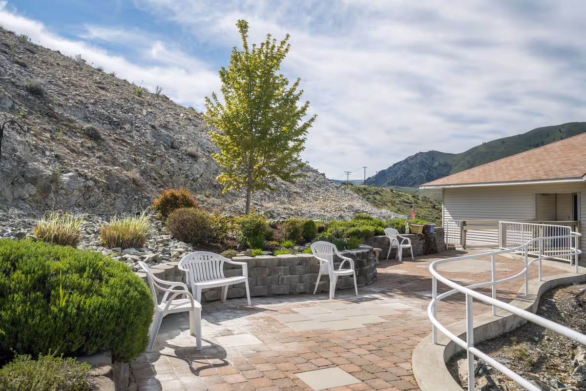 Sunlit outdoor patio with white plastic chairs around stone planters, a curved accessibility ramp, and a rocky hillside and mountains in the background.
