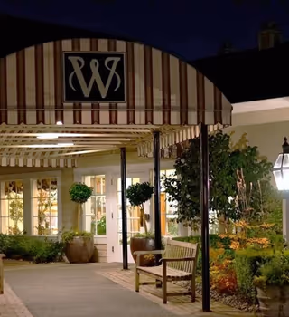 Night view of the entrance to Weatherly Inn Tacoma with a striped awning featuring a 'W' logo, a wooden bench, potted plants, and illuminated windows and outdoor lanterns.