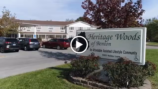 Front exterior of the Heritage Woods of Benton assisted living building with a landscaped entrance sign and parked cars in the driveway.