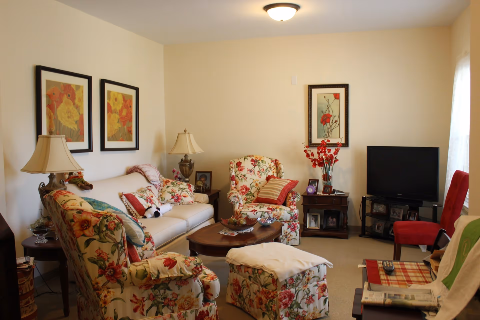 A cozy living room with floral patterned armchairs and ottoman, a white sofa with decorative pillows, two table lamps, a wooden coffee table, framed floral artwork on the walls, a flat-screen TV on a stand, and a red chair near a window with sheer curtains.