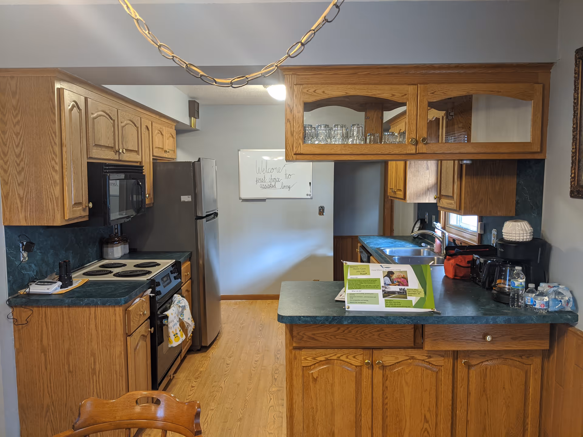 Interior view of a kitchen with wooden cabinets, a stove, a refrigerator, a sink, and a countertop with various items including a coffee maker, water bottles, and a small display stand. A whiteboard on the back wall reads 'Welcome First Choice Assisted Living'.
