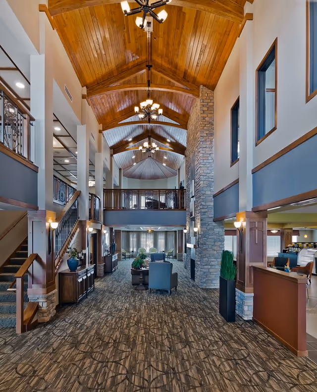 Spacious senior living facility interior with high vaulted wooden ceilings, chandeliers, stone fireplace, carpeted floor, seating area with chairs and tables, staircase with wooden railing, and large windows allowing natural light.