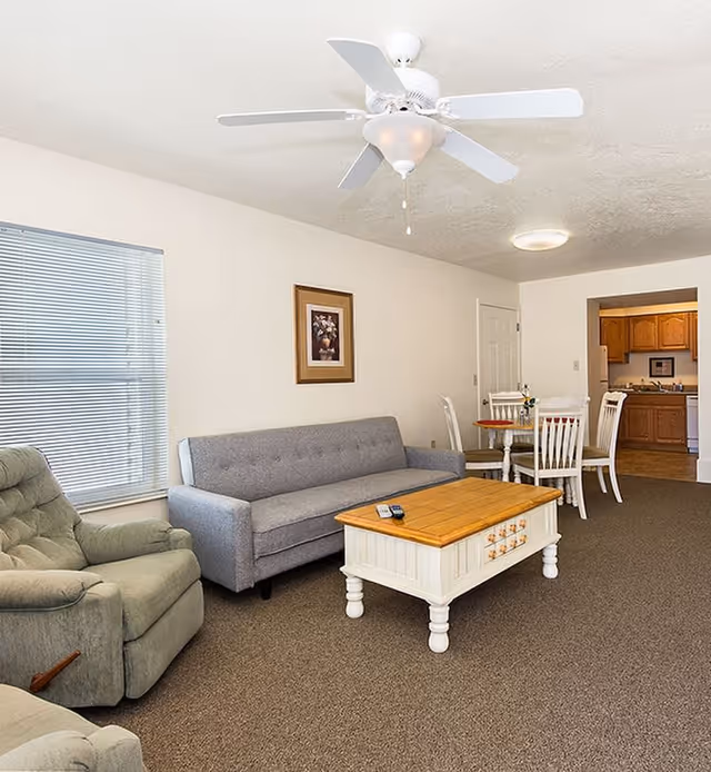 A living room area with a gray sofa, a green recliner chair, a white coffee table with a wooden top, and a ceiling fan above. In the background, there is a dining table with four white chairs and a kitchen visible through an open doorway.