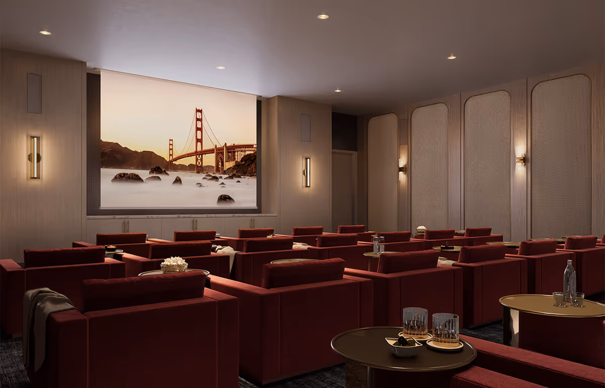 A cozy theater room with multiple rows of red upholstered armchairs facing a large screen displaying an image of the Golden Gate Bridge at sunset. Small round tables with drinks and snacks are placed between the chairs. The room has soft wall lighting and a warm, inviting atmosphere.