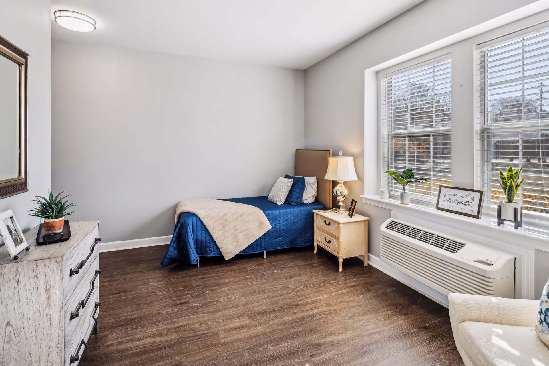 A bright and clean bedroom with a single bed covered in a blue quilt and beige throw blanket. Next to the bed is a wooden nightstand with a lamp and a small framed photo. There are two large windows with white blinds letting in natural light. A white air conditioning unit is installed below the windows. On the left side of the room is a wooden dresser with a potted plant and framed photo on top. The floor is a dark wood laminate and the walls are painted light gray.