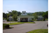 Front exterior view of a single-story building with a covered entrance and a small tower-like structure on the roof, surrounded by trees and a paved driveway.