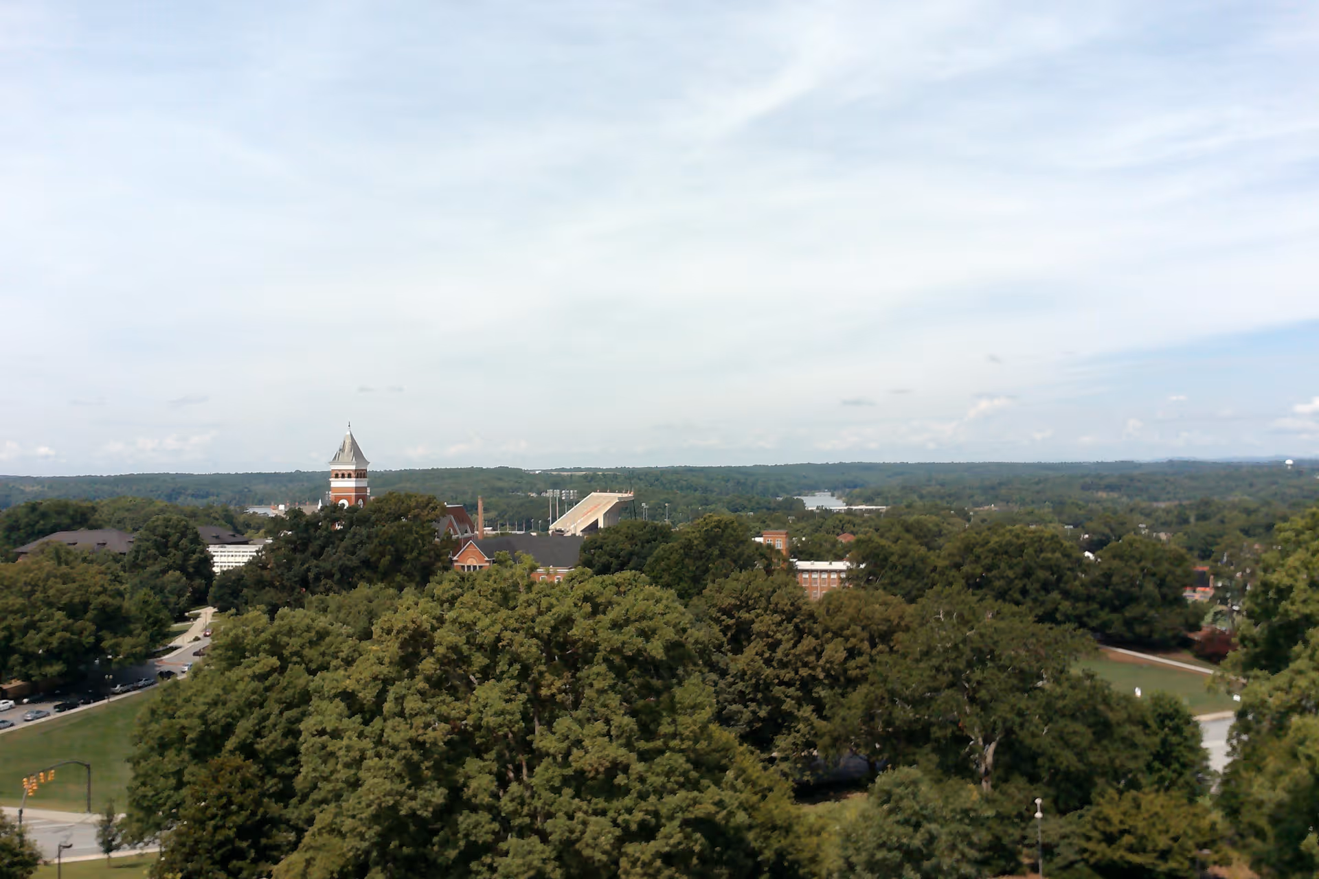 A wide aerial view of a tree-covered campus with a clock tower and stadium under a partly cloudy sky.