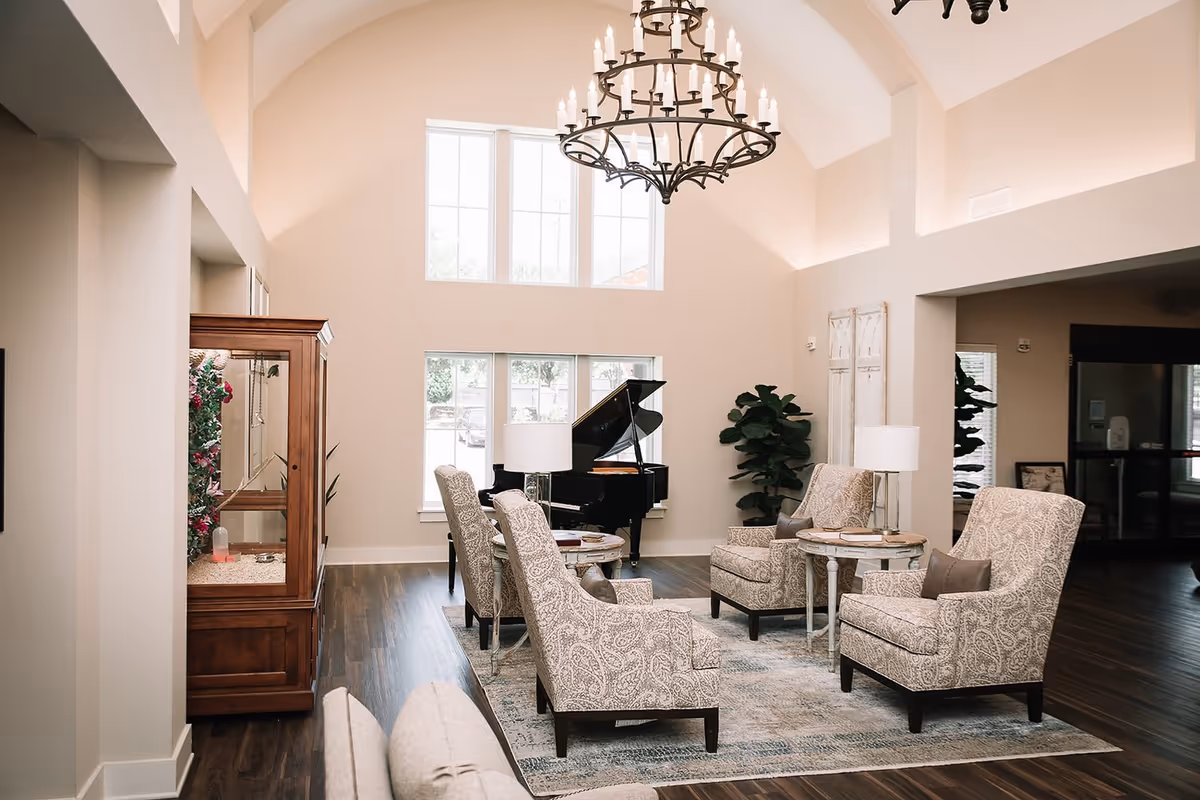 Bright high-ceiling common room with a grand piano, upholstered armchairs arranged around side tables, and a chandelier.