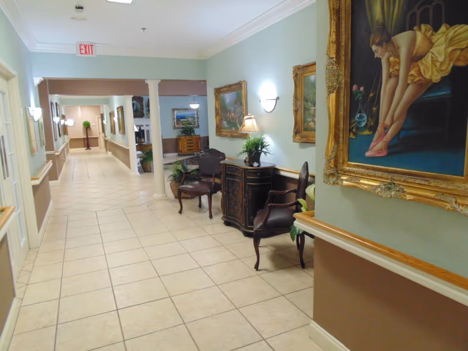 A bright tiled hallway in a senior living facility lined with framed paintings, chairs and decorative furniture.
