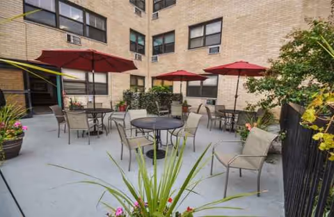 Outdoor patio area with tables, chairs, and red umbrellas beside a brick building.