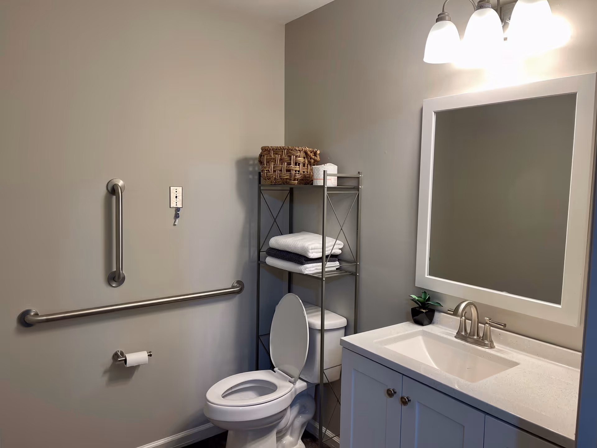 A clean bathroom with a white toilet, a metal shelving unit holding folded towels and a basket, a white sink with a faucet, a large mirror above the sink, and two grab bars mounted on the wall for accessibility.