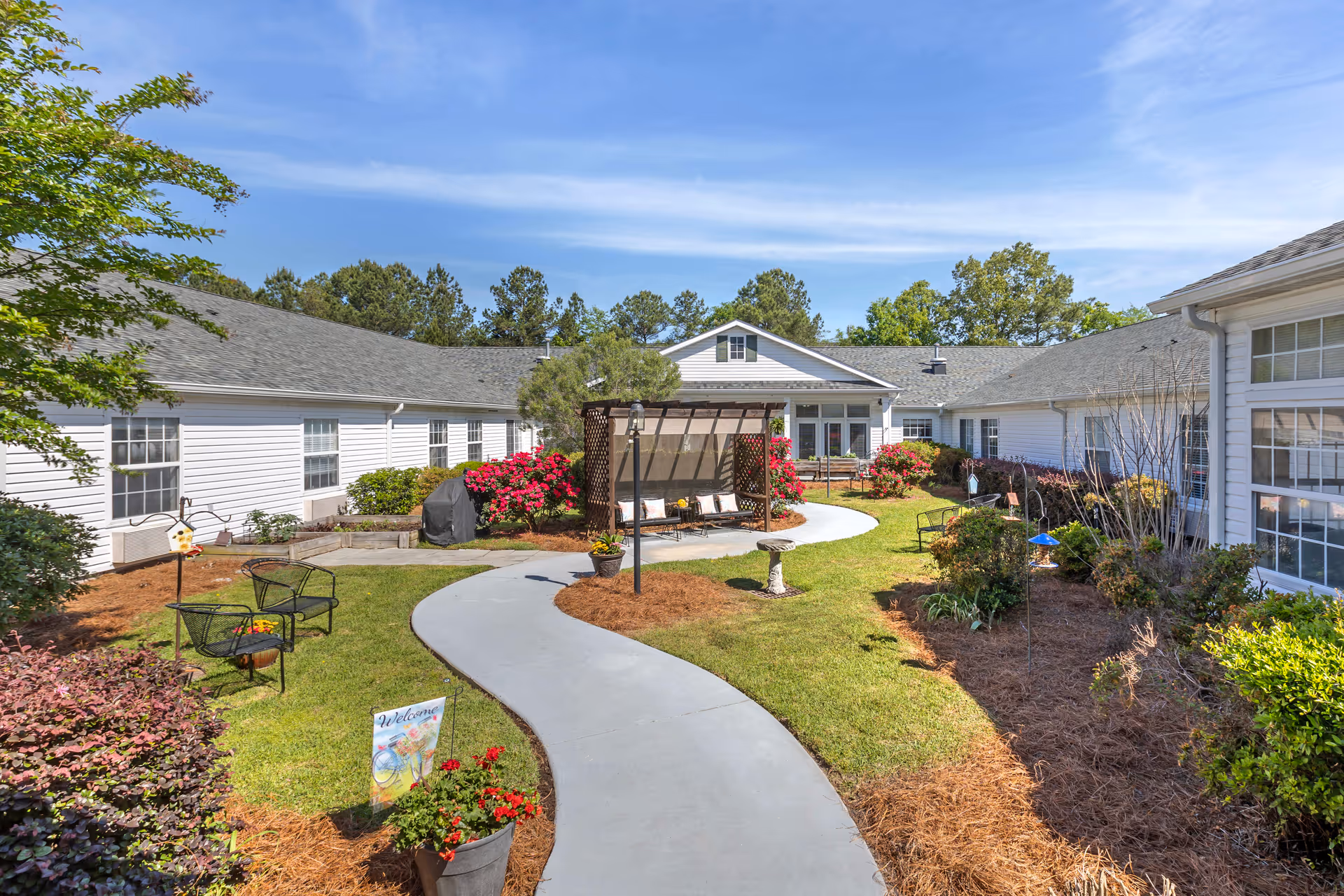 A sunny outdoor courtyard area at Brookdale Columbia featuring a winding concrete pathway, green grass, colorful flowers, shrubs, and a wooden pergola with seating underneath. The courtyard is surrounded by white buildings with multiple windows and a clear blue sky overhead.