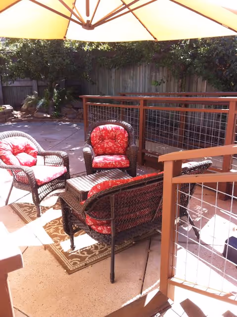 Sunny outdoor patio with wicker chairs and a loveseat featuring red floral cushions under a large umbrella beside a fenced deck.