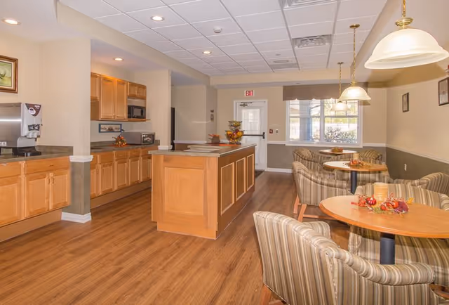 A bright and clean dining area in a senior living facility featuring wooden floors, multiple round tables with striped cushioned chairs, pendant lights hanging from the ceiling, and a kitchen area with wooden cabinets, a microwave, and a coffee machine.