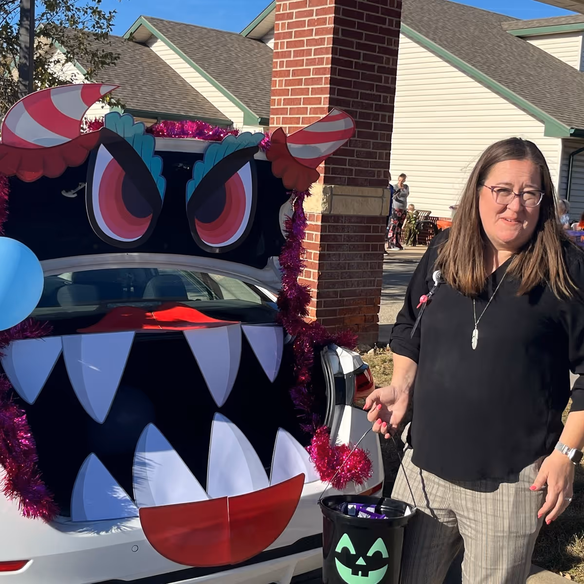 A woman stands next to a car decorated with a large monster face on its open trunk, featuring big eyes, sharp teeth, and candy cane horns. The woman is holding a black bucket with a green jack-o'-lantern face, and there are houses and other people in the background under a clear blue sky.