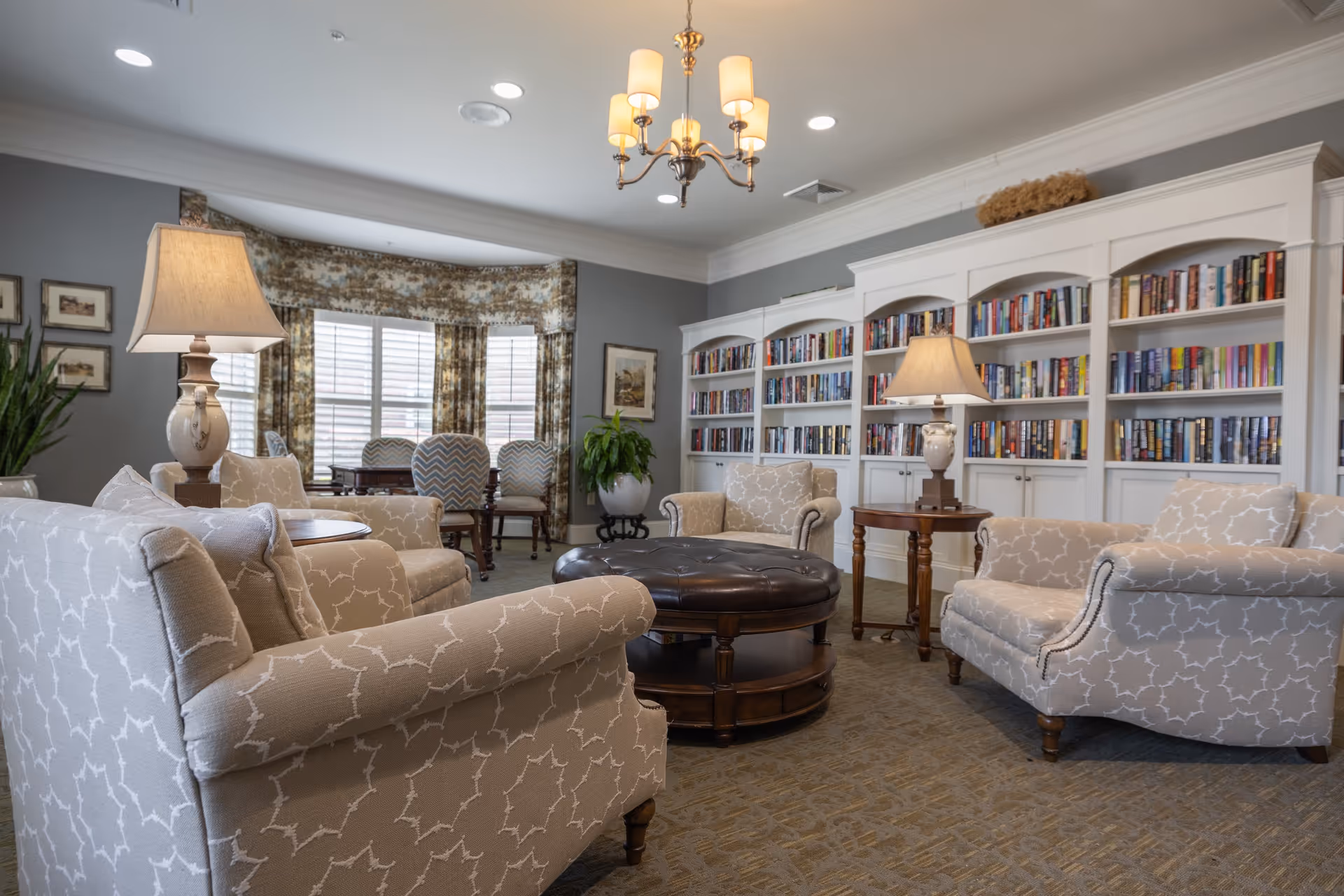 Comfortable lounge with patterned armchairs around a round leather ottoman, built-in bookshelves, table lamps, and a window with drapes.