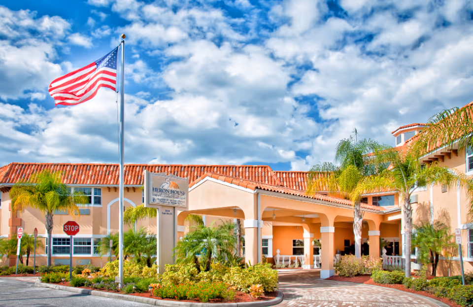 Exterior view of Heron House Assisted Living facility with a covered entrance, palm trees, landscaped bushes, an American flag on a flagpole, and a partly cloudy sky.