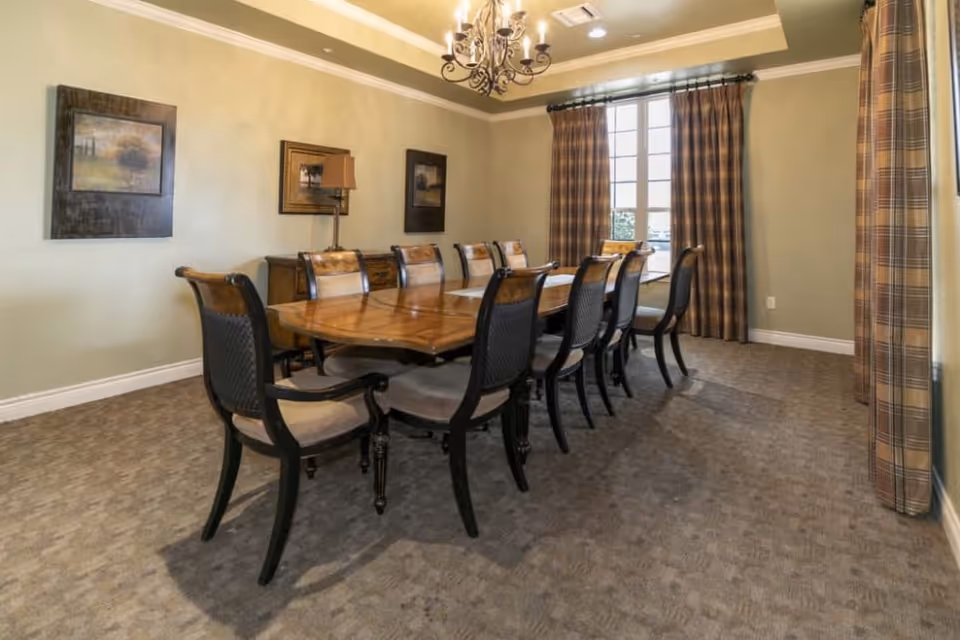 A formal dining room with a long wooden table surrounded by ten cushioned chairs. The room has beige walls adorned with framed artwork, a chandelier hanging from the ceiling, and large windows with plaid curtains allowing natural light to enter.