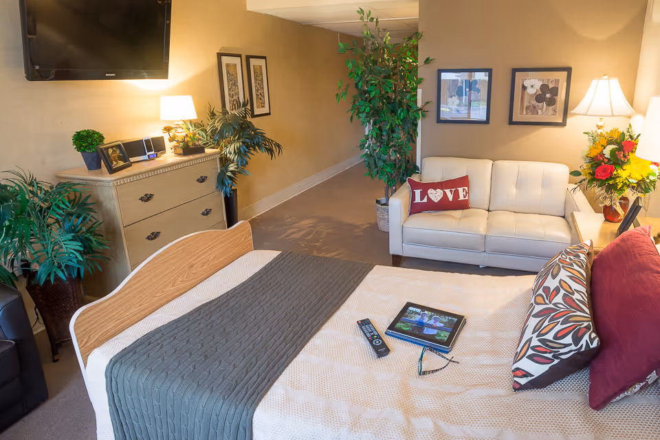 A cozy bedroom in a senior living facility featuring a bed with a gray knitted blanket and decorative pillows, a remote control and tablet on the bed, a white leather couch with a red pillow that says 'LOVE', framed artwork on the walls, a wooden dresser with plants and a lamp, and several potted plants adding greenery to the room.