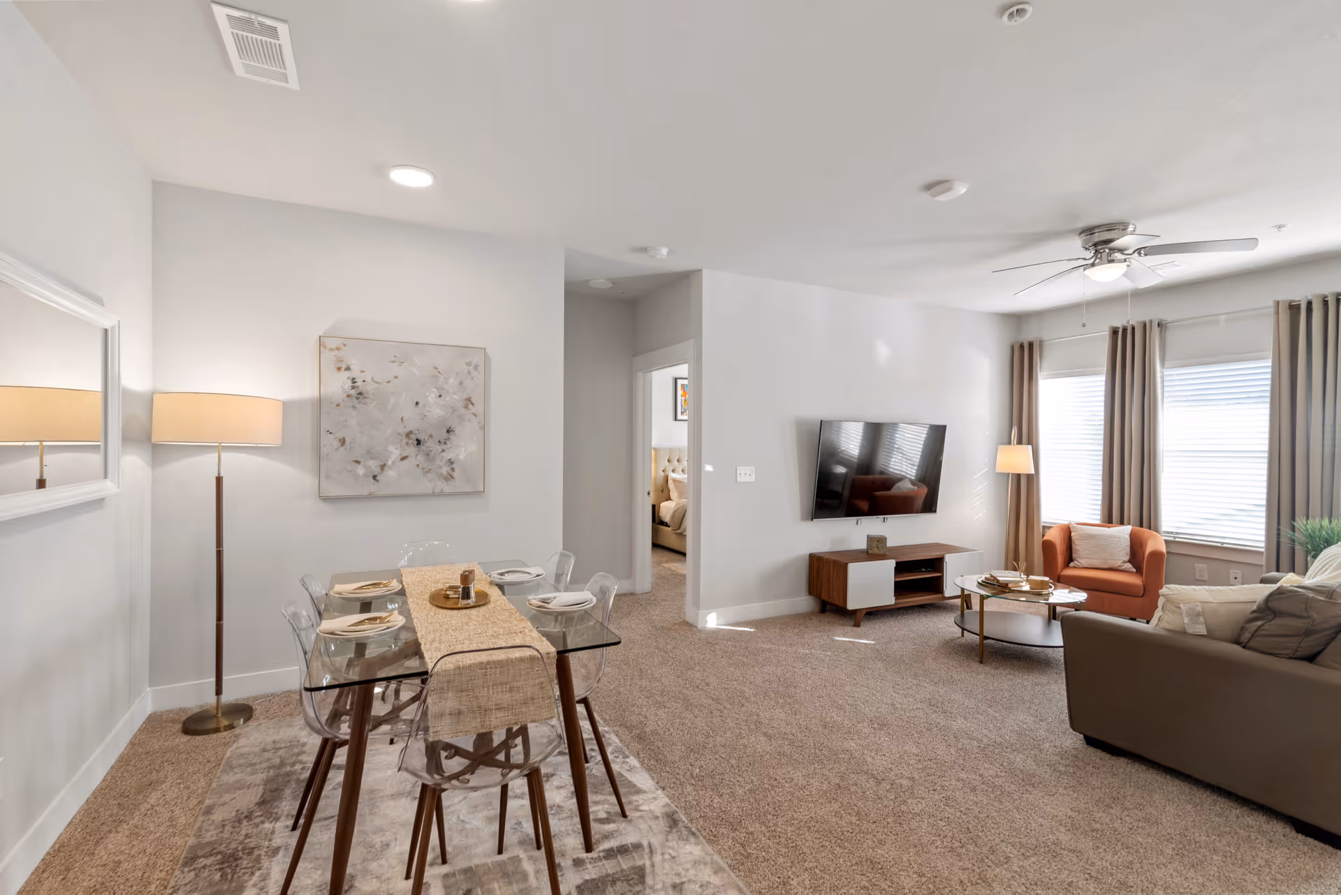 Open living room and dining area with a glass dining table, wall-mounted TV, sofa, and armchair by bright windows.