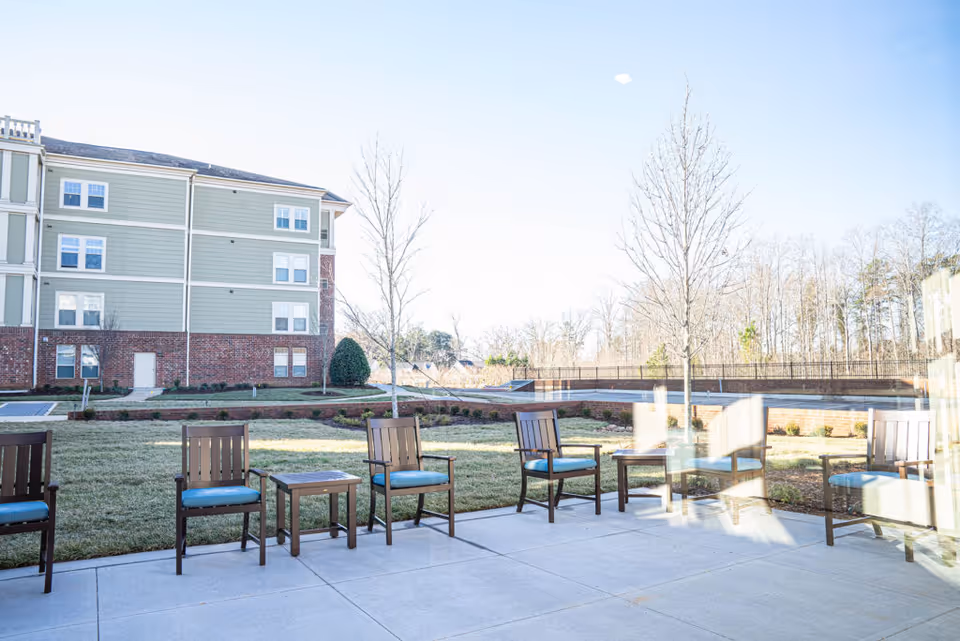 Outdoor patio area with several wooden chairs with blue cushions and small tables arranged on a concrete surface. In the background, there is a multi-story building with green siding and red brick on the lower level, along with leafless trees and a grassy lawn under a clear sky.