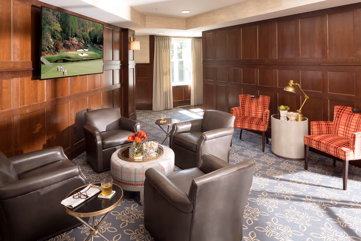A cozy sitting area with four dark leather armchairs arranged around a round ottoman with a tray holding flowers and snacks. Two red plaid upholstered chairs are placed against a wood-paneled wall with a brass lamp and small table between them. A flat-screen TV is mounted on the wood-paneled wall showing a golf course scene. The room has a patterned carpet and a window with beige curtains letting in natural light.