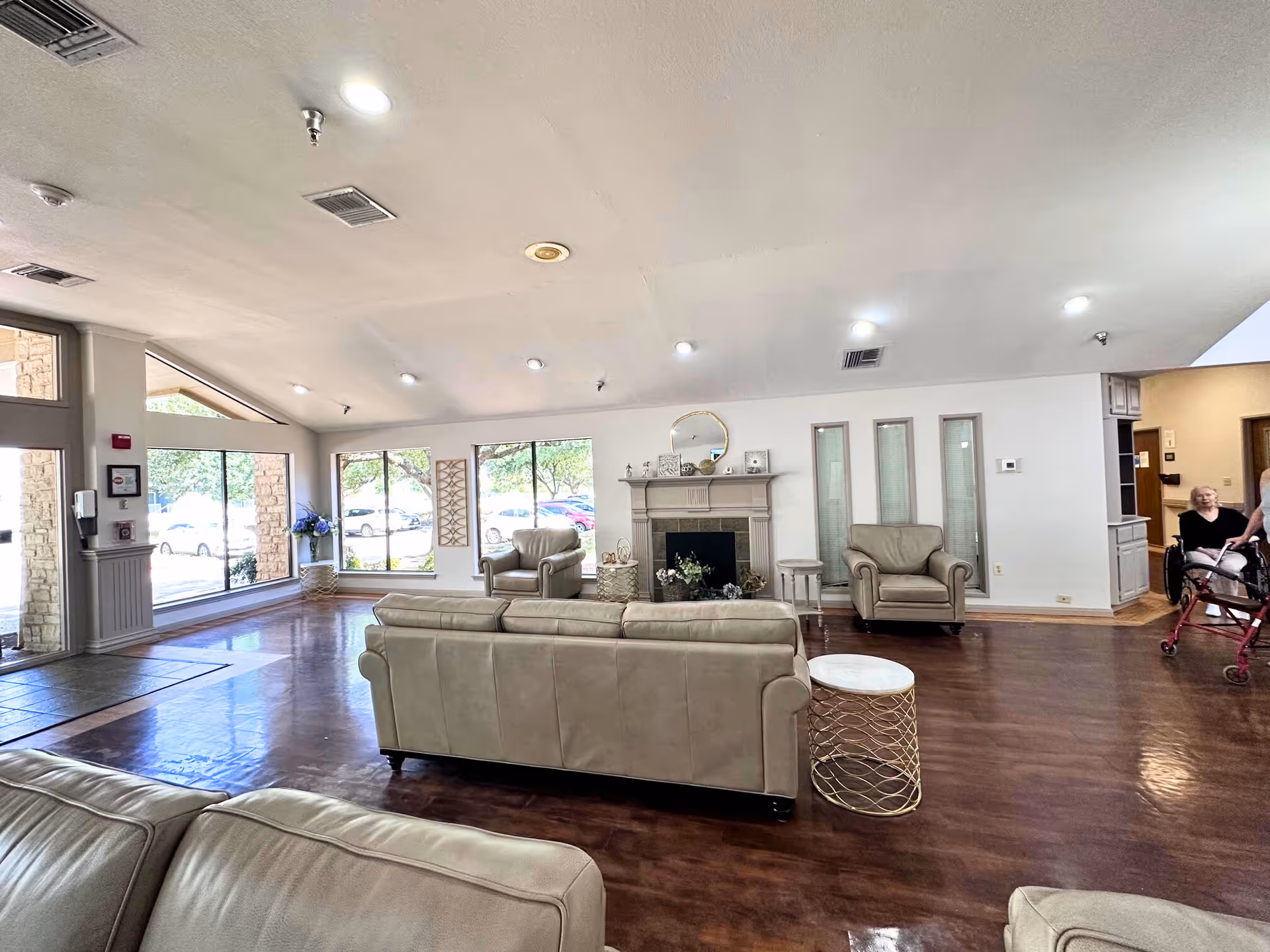 Bright common living room with leather sofas and armchairs centered around a fireplace, large windows along one wall, and two residents near the doorway.