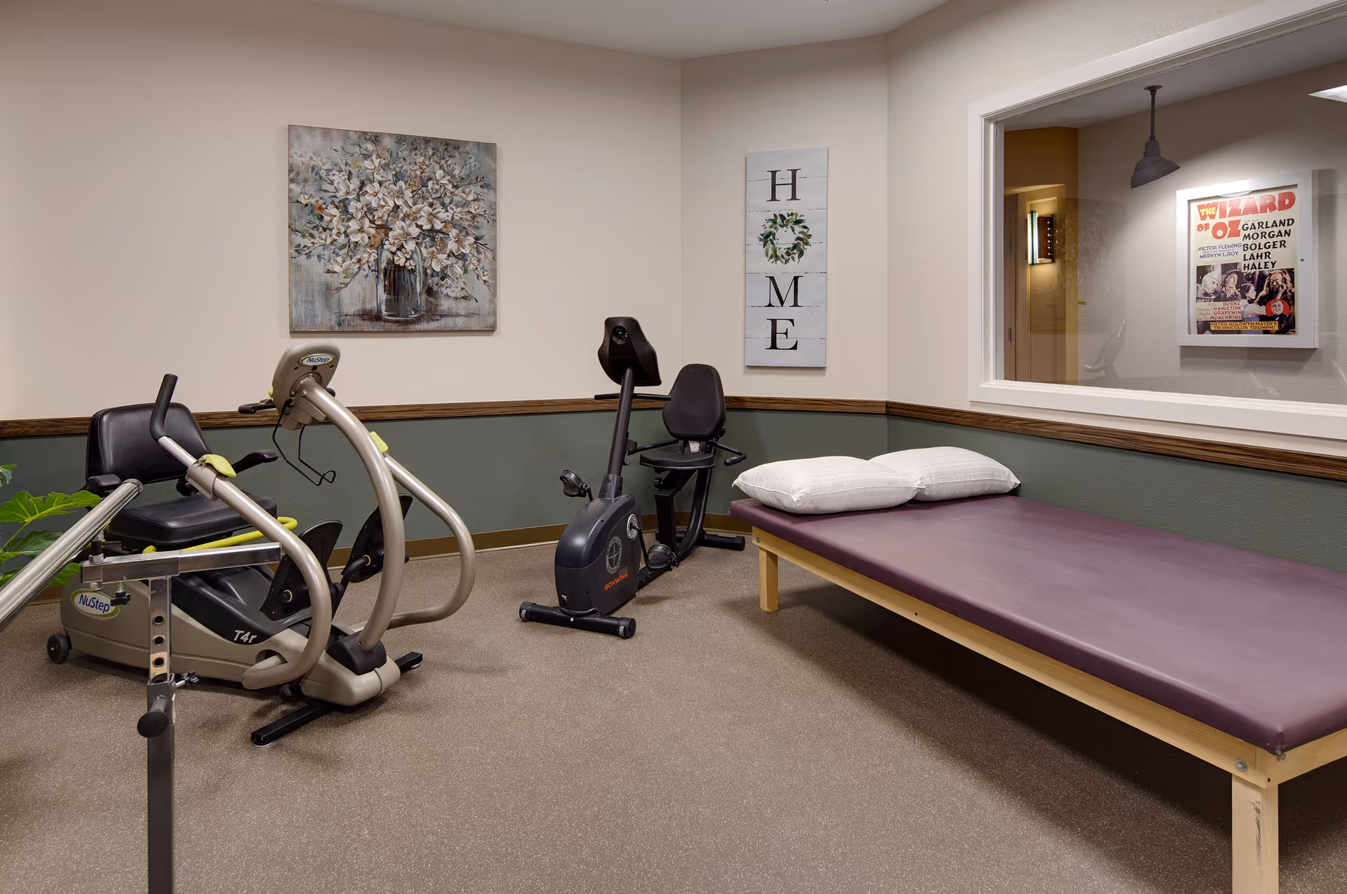 Therapy/exercise room with two recumbent stationary bikes, a padded treatment table with pillows, and wall decor.