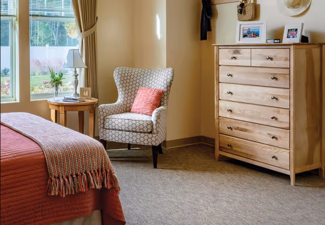A cozy bedroom corner with a patterned armchair featuring a red cushion, a wooden dresser with framed photos on top, a round wooden side table with a lamp and a small plant, and a bed with a red quilt and a striped throw blanket. Sunlight streams through a window with beige curtains.