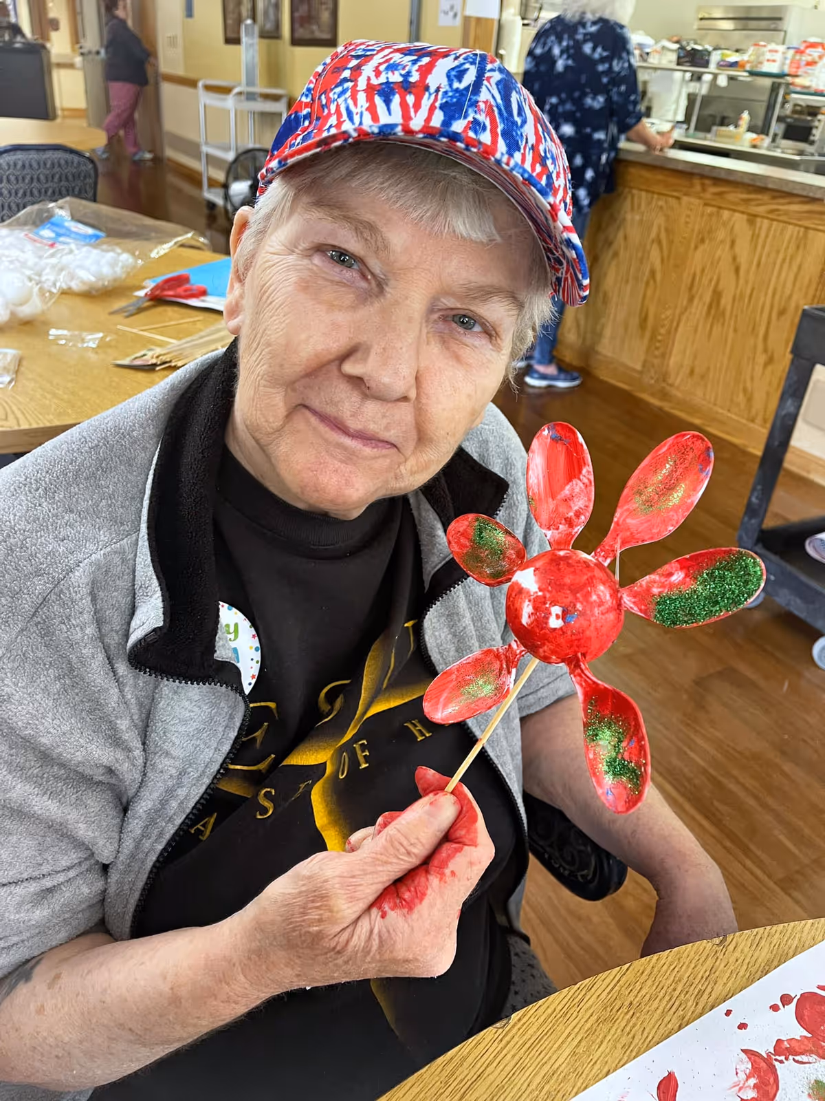 An elderly woman wearing a red, white, and blue patterned cap and a gray jacket sits at a table in a communal room. She is holding a red and green painted craft shaped like a flower made from spoons. There are art supplies and other people in the background.