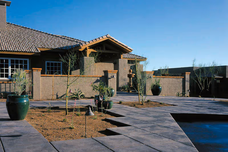 Outdoor patio area with a tiled walkway, several large green ceramic pots with plants, and a beige stucco building with a tiled roof in the background under a clear blue sky.