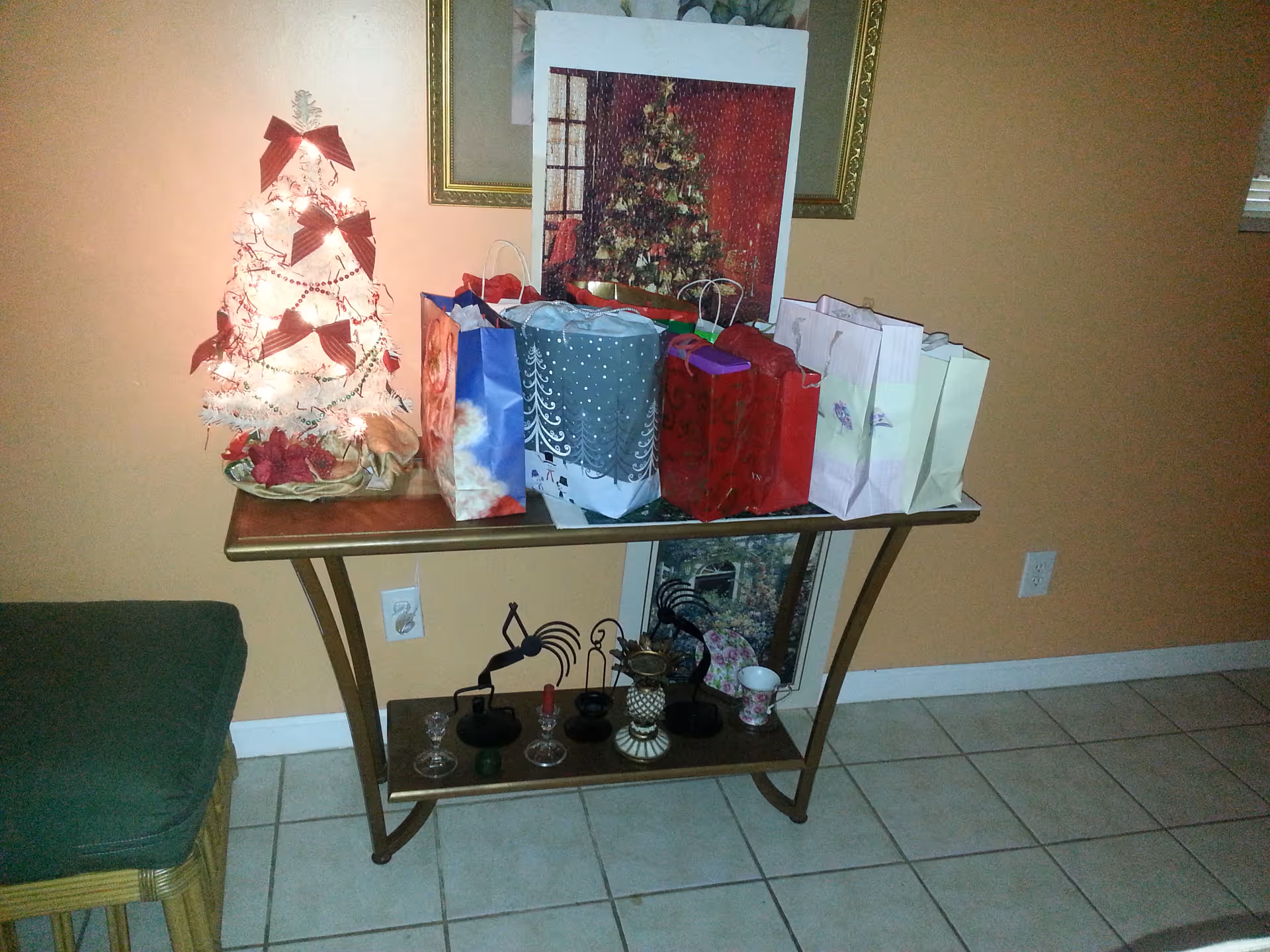 A small table against a beige wall with a lit white Christmas tree decorated with red bows and beads. Several colorful gift bags are arranged on the table. Below the table are decorative items including candle holders and a small figurine. A green cushioned stool is partially visible on the left side.