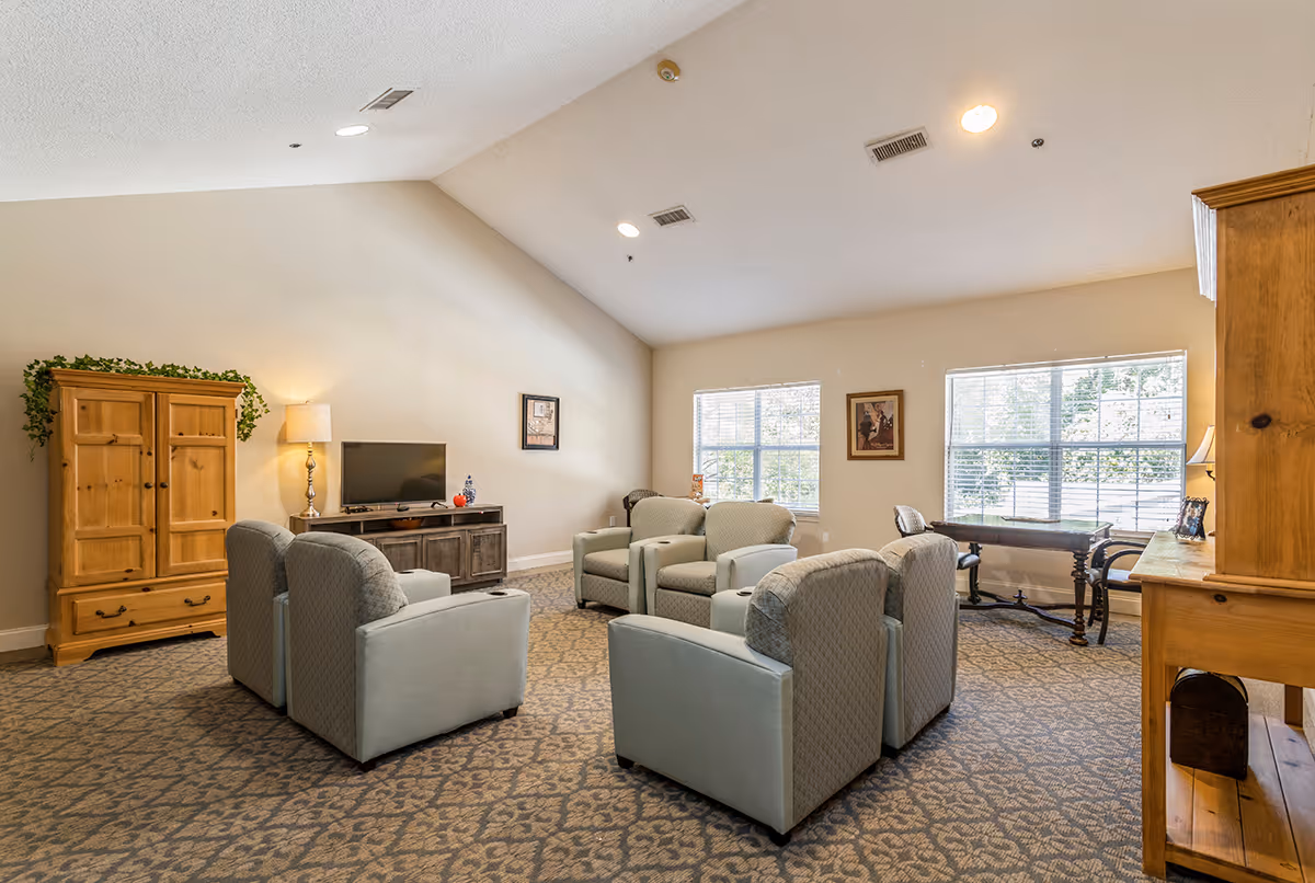 A cozy living room area with six cushioned armchairs arranged in a circle on a patterned carpet. The room features a wooden TV cabinet with a flat-screen TV, a wooden armoire with a plant on top, two large windows letting in natural light, framed artwork on the walls, and a wooden desk with a chair near the windows.