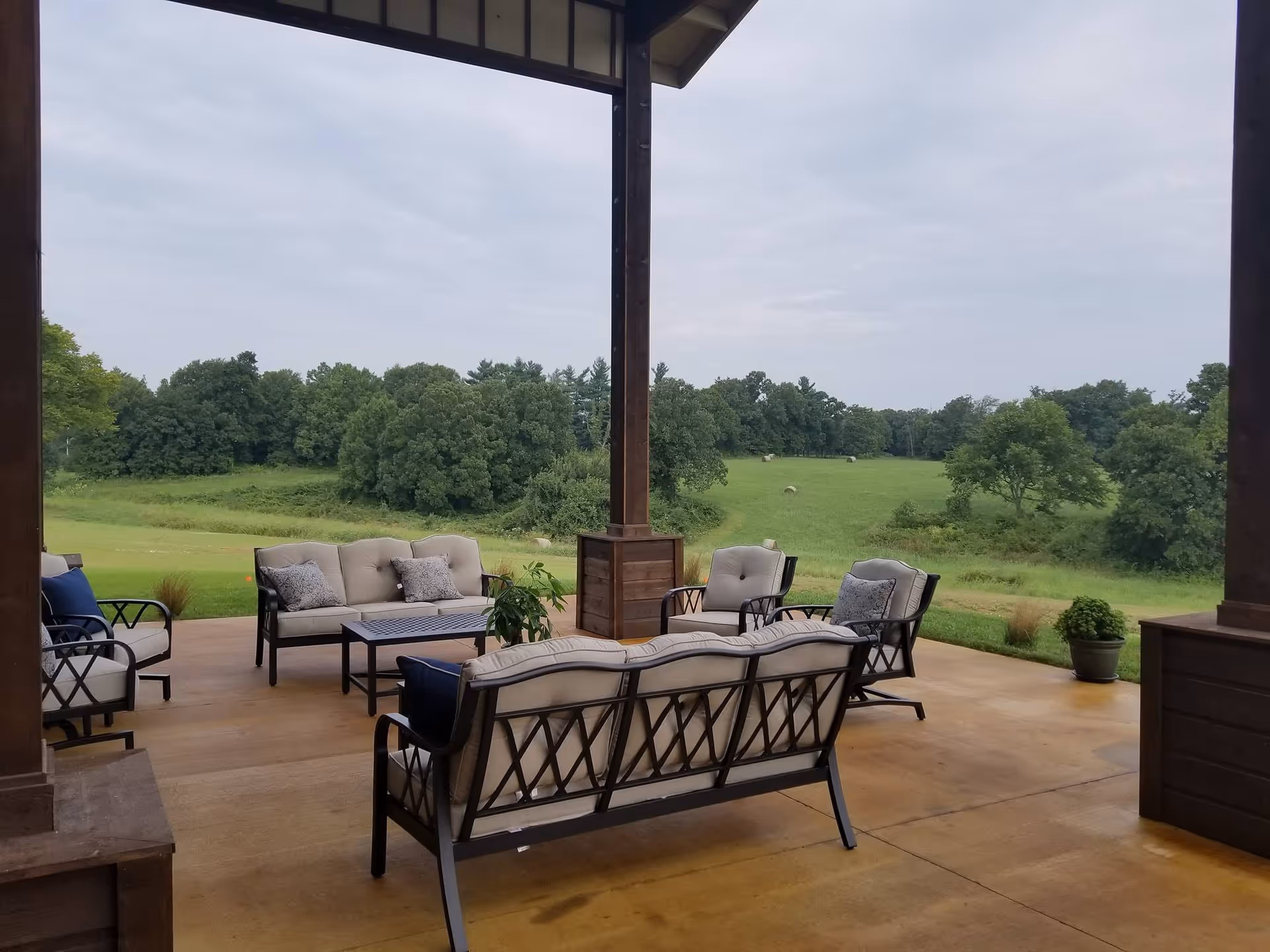 Outdoor covered patio area with cushioned metal chairs and sofas arranged around a coffee table, overlooking a green field with trees and a cloudy sky.