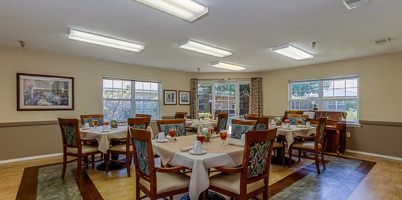 Bright dining room with multiple round tables set with tablecloths, chairs, and glasses in a senior living facility.