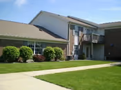 Exterior view of a two-story senior living facility building with brick and siding walls, several windows, a small balcony, and a well-maintained lawn with bushes and a concrete walkway.