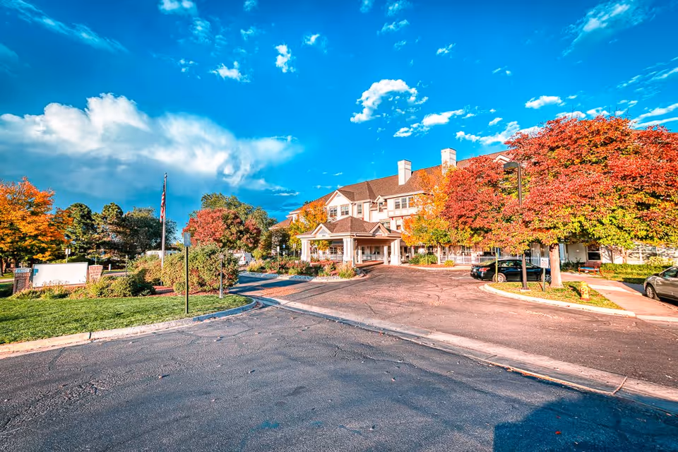 Exterior view of Woodland Estates Senior Living facility with a clear blue sky, colorful autumn trees with red and orange leaves, a driveway leading to the entrance, and parked cars near the building.