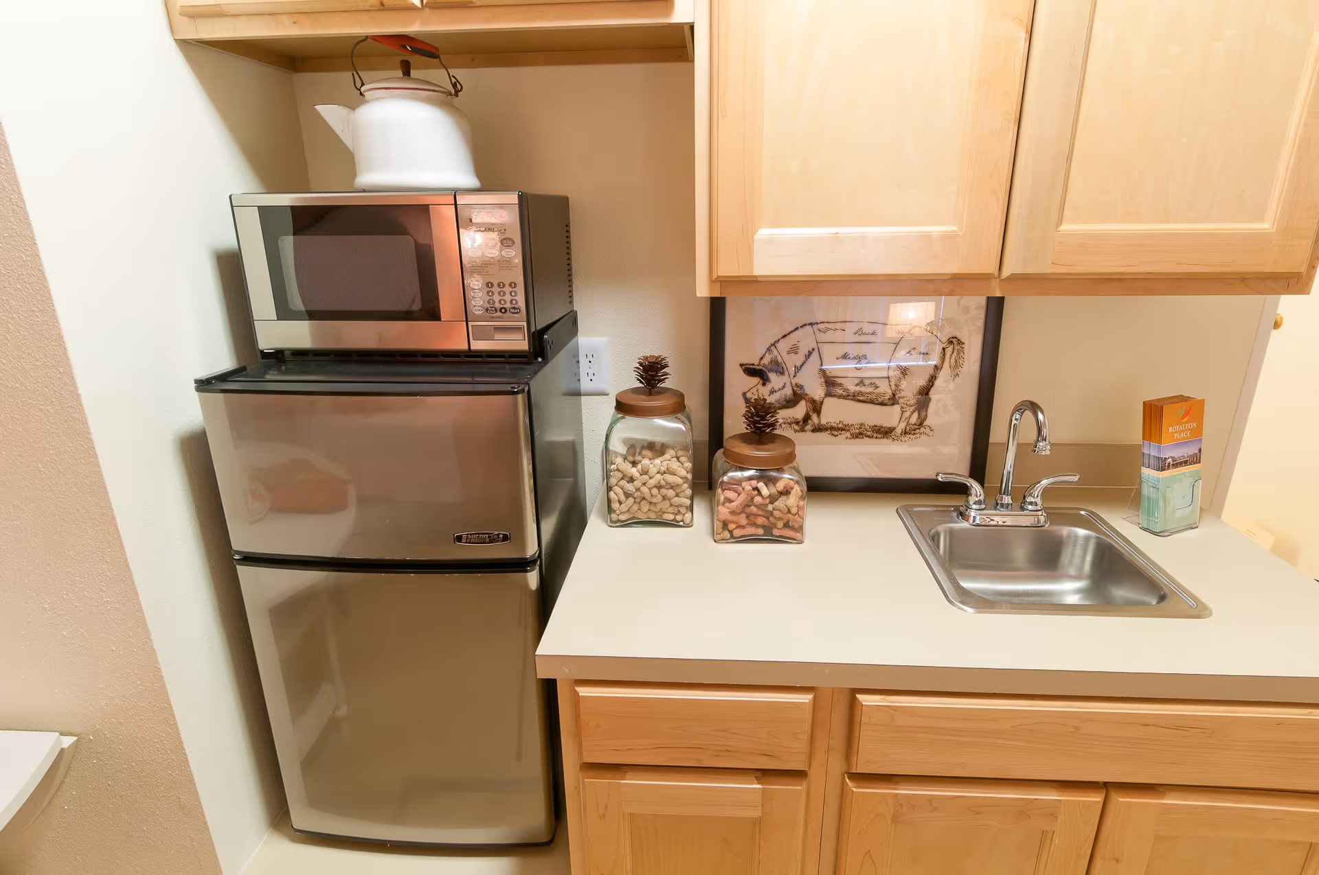 A small kitchen area with a stainless steel mini refrigerator and microwave stacked on top. There is a white kettle on top of the microwave. Next to the refrigerator is a countertop with a small stainless steel sink and faucet. On the counter are two glass jars filled with dog treats and a framed picture of a pig diagram. A brochure or pamphlet labeled Royalton Place is also on the counter. Light wood cabinets are mounted above and below the countertop.