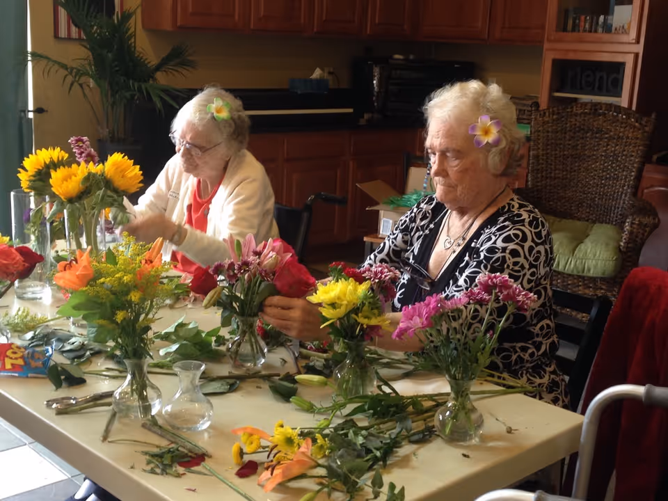 Two elderly women sitting at a table indoors, arranging colorful flowers in small glass vases. Both women have flowers tucked behind their ears. The table is covered with various flowers and leaves. The background shows wooden cabinets and a wicker chair with a green cushion.