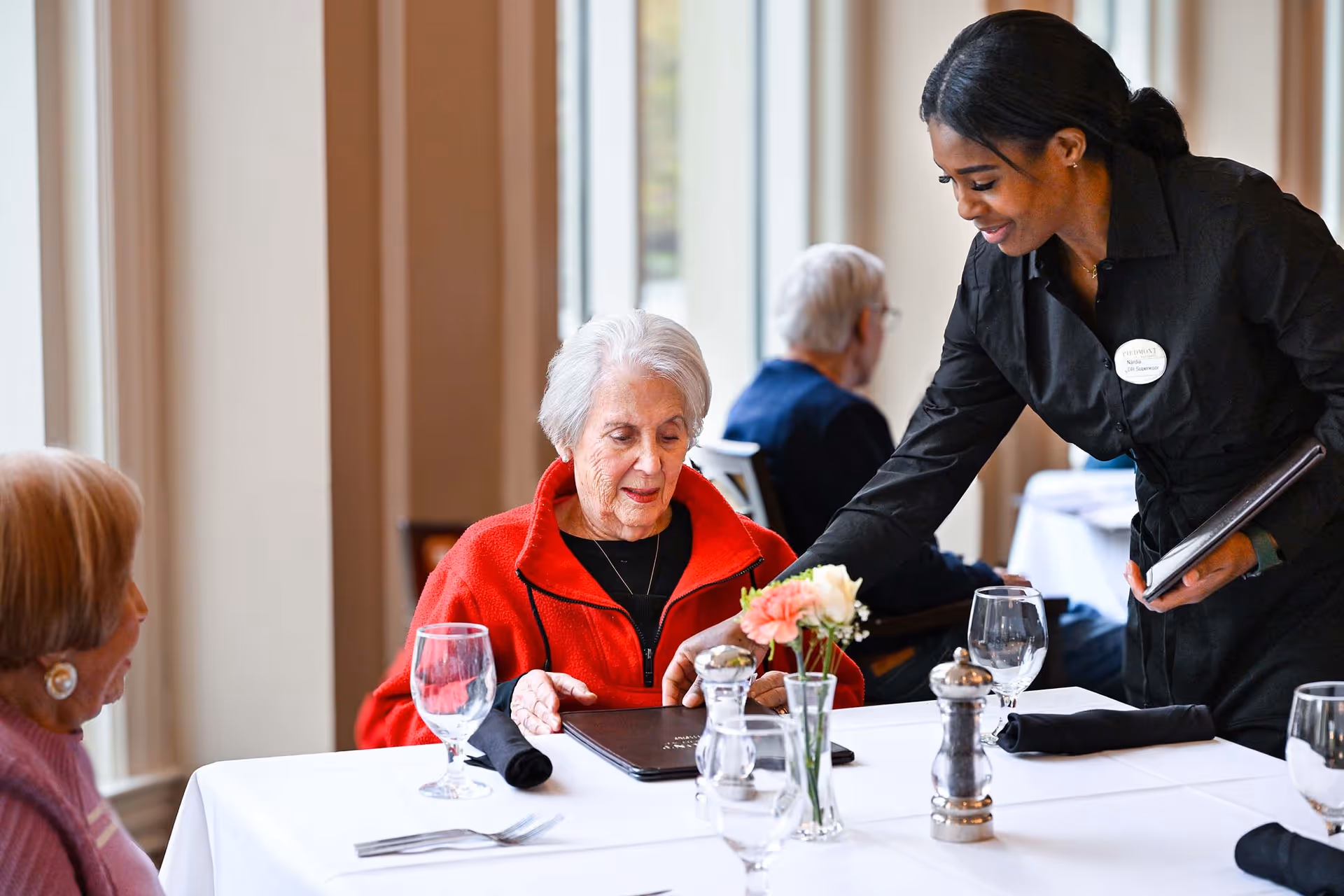 A waitress in a black uniform is serving two elderly women seated at a dining table with white tablecloth, glassware, and a small flower vase in a well-lit dining room.