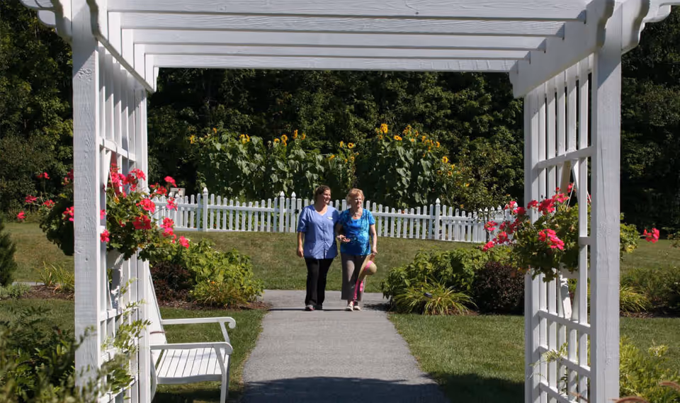 Two women walking together on a paved garden path under a white wooden pergola with hanging pink flowers. The path is surrounded by green grass, bushes, and sunflowers in the background, with a white picket fence behind them.