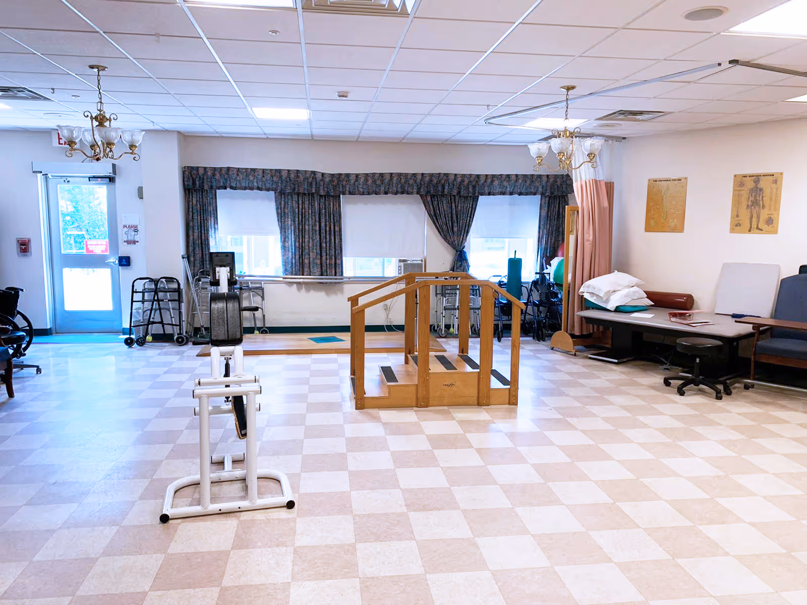 A rehabilitation room with exercise equipment including a stationary bike and a wooden set of stairs with handrails for physical therapy. The room has checkered tile flooring, large windows with floral curtains, and various medical and therapy supplies such as walkers and wheelchairs near the walls. There are also anatomical charts on the wall and a treatment table with pillows and cushions.