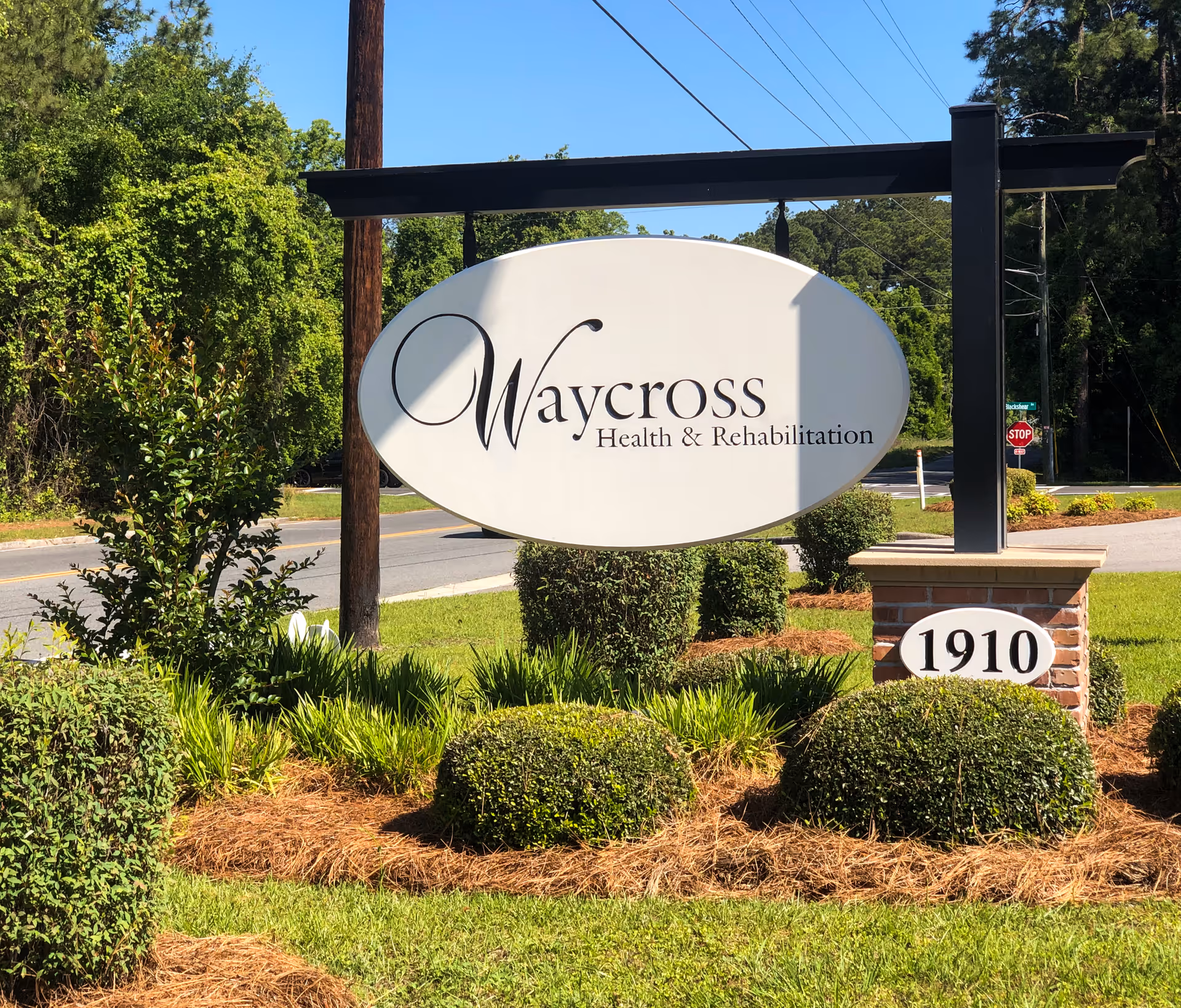 An oval sign reading 'Waycross Health & Rehabilitation' with an address plaque '1910' set among trimmed shrubs by a roadside.