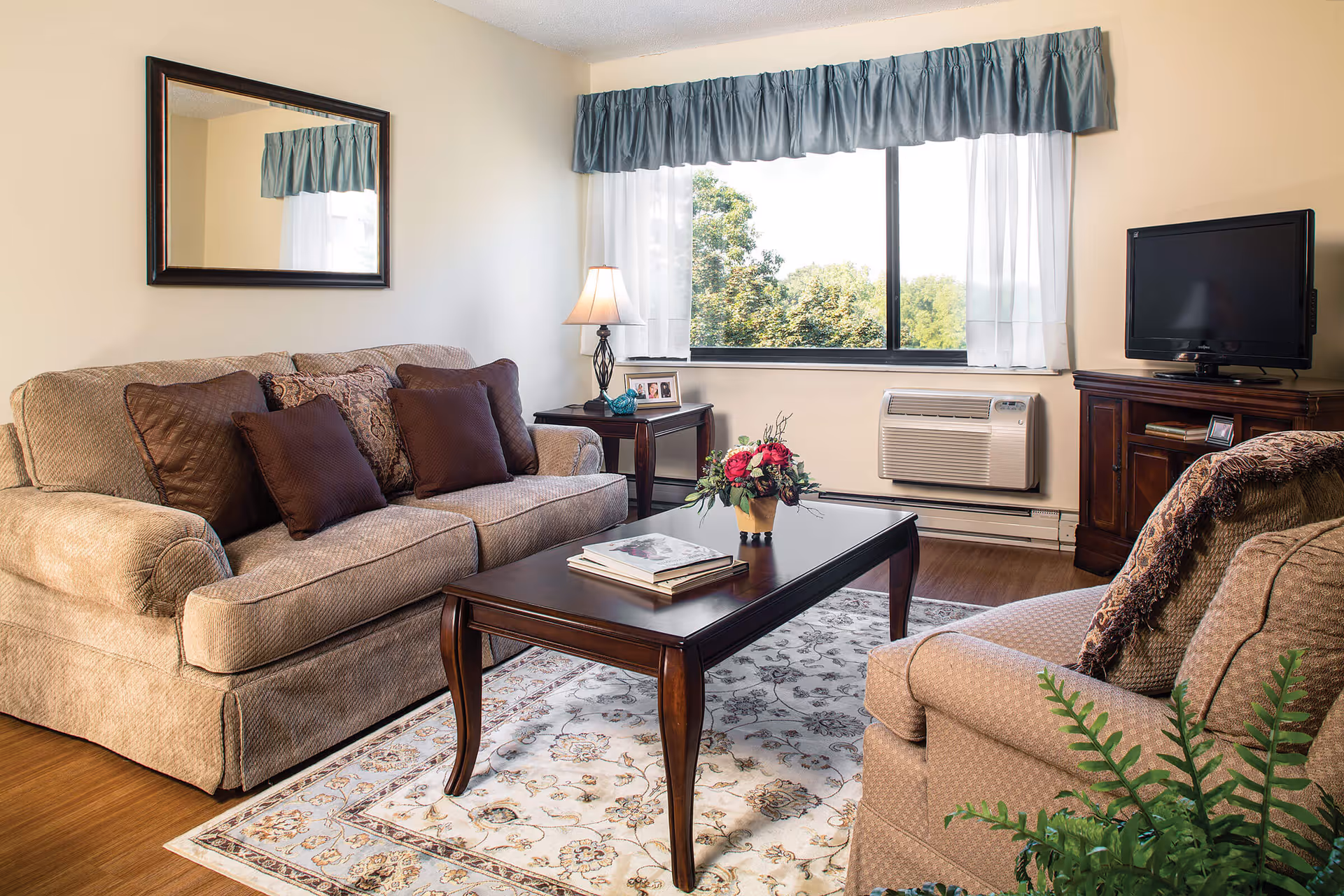 Sunlit living room with two upholstered sofas, a wooden coffee table topped with a floral arrangement, a TV, and a large window with a valance.